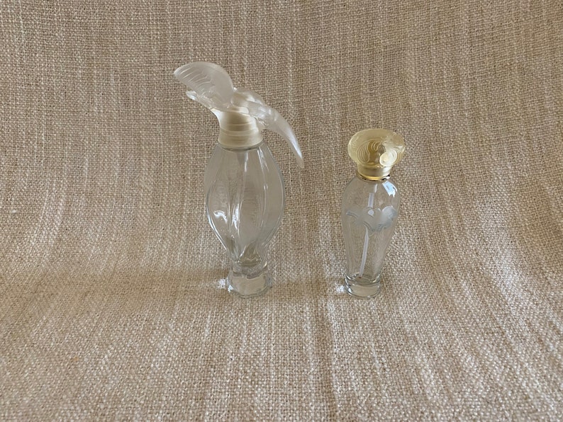 May include: Two clear glass perfume bottles with different shaped stoppers. The larger bottle has a clear, sculpted stopper shaped like a bird. The smaller bottle has a gold-colored, round stopper.
