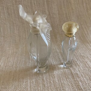 May include: Two clear glass perfume bottles with decorative stoppers. The bottle on the left has a bird-shaped stopper, while the bottle on the right has a floral-shaped stopper.