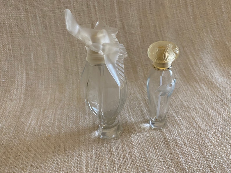 May include: Two clear glass perfume bottles with decorative stoppers. The bottle on the left has a bird-shaped stopper, while the bottle on the right has a floral-shaped stopper.