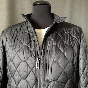 Könnte beinhalten: Eine schwarze Steppjacke mit Rautenmuster, Stehkragen und einer vertikalen Reißverschlusstasche auf der Brust. Die Jacke verfügt über einen zentralen Frontreißverschluss mit metallischen Elementen und wird auf einer Schaufensterpuppe gezeigt.