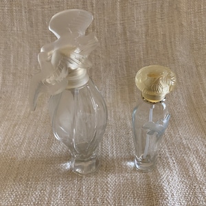 May include: Two clear glass perfume bottles with decorative stoppers. The larger bottle has a white bird-shaped stopper. The smaller bottle has a yellow floral stopper.