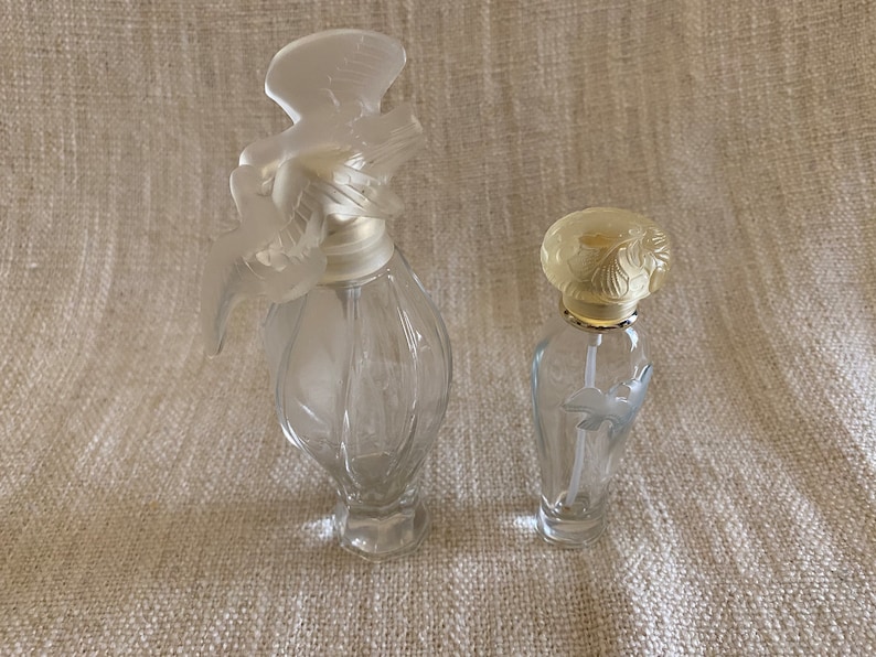 May include: Two clear glass perfume bottles with decorative stoppers. The larger bottle has a white bird-shaped stopper. The smaller bottle has a yellow floral stopper.