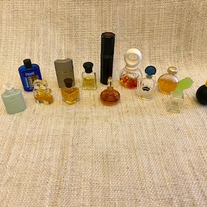 Könnte beinhalten: Eine Sammlung von Miniatur-Parfümfläschchen und eine kleine Flasche Duschgel. Die Flaschen sind in einer Reihe auf einer beigefarbenen, strukturierten Oberfläche angeordnet. Die Flaschen sind in verschiedenen Farben, darunter Blau, Grün, Gold und Schwarz.