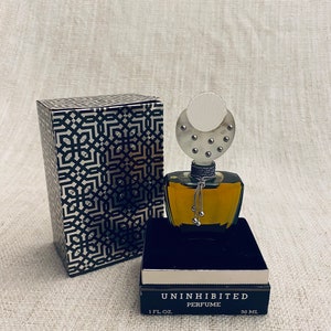 Puede incluir: Una caja negra y plateada con un patr&oacute;n geom&eacute;trico y una botella de perfume negra con una tapa plateada y una cadena plateada con cascabeles. La botella est&aacute; etiquetada como "Uninhibited Perfume" y "1 fl. oz. 30 ml."