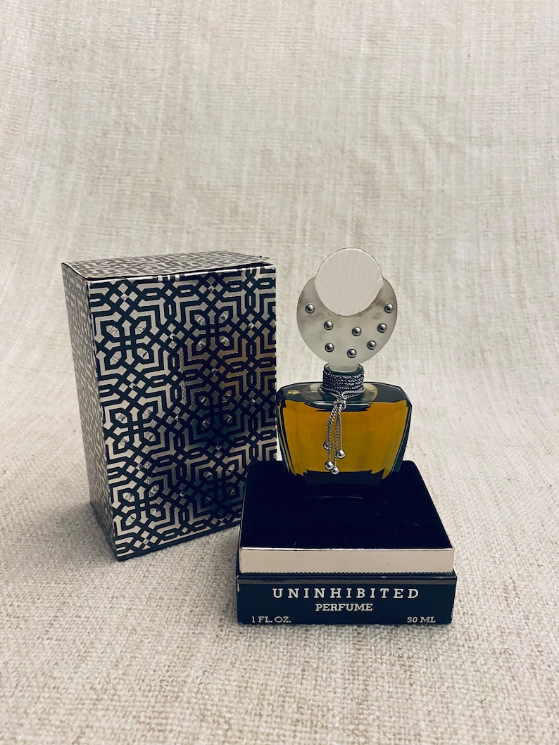 Puede incluir: Una caja negra y plateada con un patr&oacute;n geom&eacute;trico y una botella de perfume negra con una tapa plateada y una cadena plateada con cascabeles. La botella est&aacute; etiquetada como "Uninhibited Perfume" y "1 fl. oz. 30 ml."