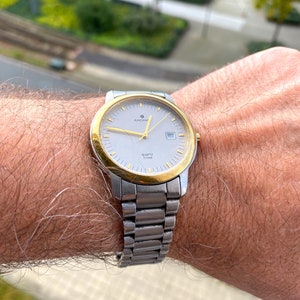 Puede incluir: Un reloj de pulsera de dos tonos, plateado y dorado, con una esfera blanca y agujas negras. La esfera del reloj tiene las palabras "Juncheng" y "Quartz Titan" impresas.