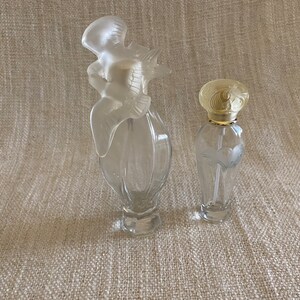 May include: Two clear glass perfume bottles with decorative stoppers. The larger bottle has a bird-shaped stopper and the smaller bottle has a floral design stopper.