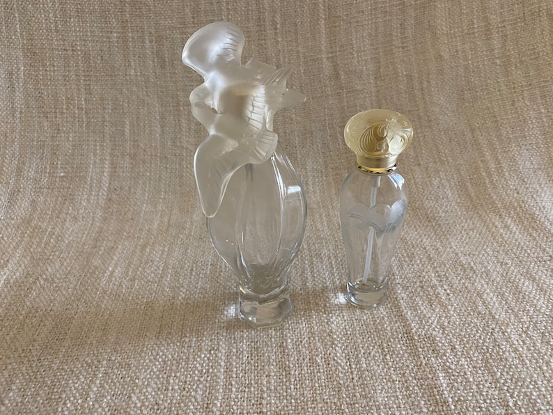 May include: Two clear glass perfume bottles with decorative stoppers. The larger bottle has a bird-shaped stopper and the smaller bottle has a floral design stopper.