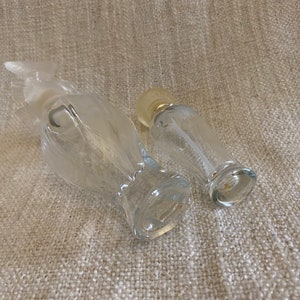 May include: Two clear glass perfume bottles, one shaped like a bird and the other a cylinder. Both bottles have a silver top.