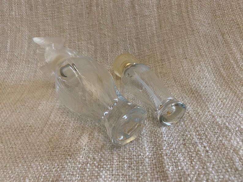 May include: Two clear glass perfume bottles, one shaped like a bird and the other a cylinder. Both bottles have a silver top.