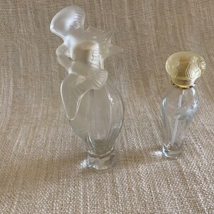 May include: Two clear glass perfume bottles with decorative stoppers. The larger bottle has a bird-shaped stopper, while the smaller bottle has a floral design stopper.