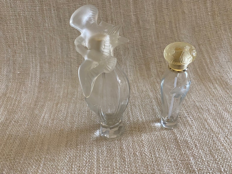 May include: Two clear glass perfume bottles with decorative stoppers. The larger bottle has a bird-shaped stopper, while the smaller bottle has a floral design stopper.