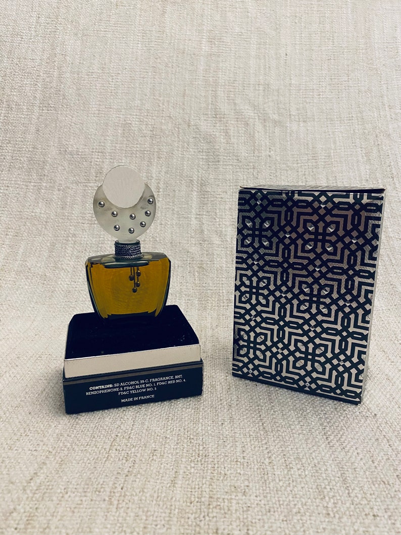 Puede incluir: Un frasco de perfume negro y plateado con una tapa plateada y un patr&oacute;n geom&eacute;trico en la caja. El frasco contiene un l&iacute;quido transparente y la caja tiene el texto "CONTAINS 50 ALCOHOL 35-C. FRAGRANCE MIT RENZOFUENONE - FD&C BLUE NO. 1 FD&C RED NO. 4 FD&C YELLOW NO. 1 MADE IN FRANCE".