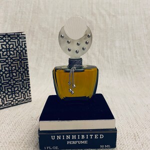 Puede incluir: Un frasco de perfume de vidrio transparente con un tap&oacute;n en forma de media luna de plata. El frasco est&aacute; sentado en una caja de terciopelo negro. La caja est&aacute; etiquetada "Uninhibited Perfume 1 fl. oz. 30 ml."