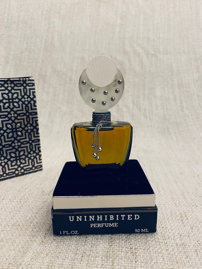 Puede incluir: Un frasco de perfume de vidrio transparente con un tap&oacute;n en forma de media luna de plata. El frasco est&aacute; sentado en una caja de terciopelo negro. La caja est&aacute; etiquetada "Uninhibited Perfume 1 fl. oz. 30 ml."