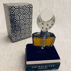 Puede incluir: Un frasco de perfume negro y dorado con una tapa plateada y una cadena plateada con cuentas que cuelgan de ella. El frasco est&aacute; sentado en una caja negra con las palabras "Uninhibited Perfume 1 fl. oz. 30 ml" impresas en ella.