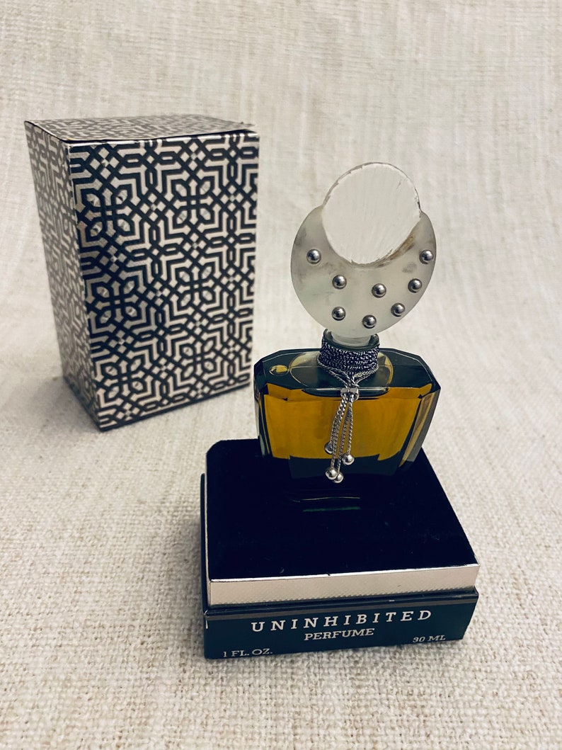 Puede incluir: Un frasco de perfume negro y dorado con una tapa plateada y una cadena plateada con cuentas que cuelgan de ella. El frasco est&aacute; sentado en una caja negra con las palabras "Uninhibited Perfume 1 fl. oz. 30 ml" impresas en ella.