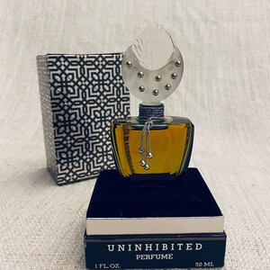 Puede incluir: Un frasco de perfume Uninhibited en un frasco de vidrio transparente con una tapa plateada y una cadena plateada con cuentas plateadas que cuelgan de la tapa. El frasco est&aacute; sentado en una caja negra con un borde plateado. La caja tiene las palabras "Uninhibited Perfume" y "1 fl. oz. 30 ml" impresas en ella.