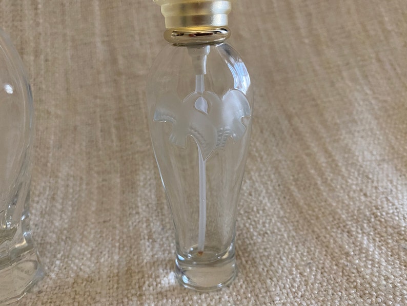 May include: A clear glass bottle with a silver top and a white floral design. The bottle is empty and has a small opening at the top.