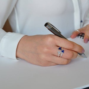 May include: A person's hand wearing a silver ring with blue triangular stones, holding a silver pen and writing in a notebook.