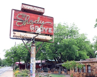 Puede incluir: Un letrero rojo y blanco con el texto "Hamburgers" y "Shady Grove" en letras de neón blancas. El letrero está montado en un poste de madera con una flecha blanca que apunta hacia la derecha. El letrero está frente a un césped verde con un edificio de piedra en el fondo.