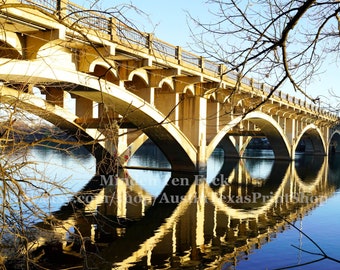 May include: A concrete bridge with multiple arches spans a body of water. The bridge is reflected in the water, creating a symmetrical pattern. The water is a deep blue color, and the sky is a bright blue.