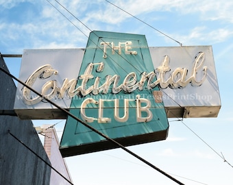 Puede incluir: Un letrero de neón vintage para "The Continental Club" con letras blancas sobre un fondo azul verdoso. El letrero está colgado de cables y tiene un aspecto descolorido y desgastado.