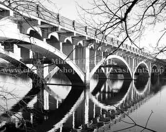 May include: A black and white photograph of a concrete bridge with multiple arches over a still body of water. The reflection of the bridge is visible in the water.