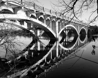 May include: A black and white photograph of a concrete bridge with multiple arches over a still body of water. The bridge is reflected in the water.