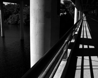 May include: A black and white photo of a concrete bridge overpass with a metal railing and shadows on the ground.