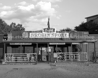 May include: Black and white photograph of the Broken Spoke, a rustic building with a wooden facade. The sign reads "Broken Spoke" above the entrance. Decorative wagon wheels adorn the exterior. A Coca-Cola sign and a flag are visible.