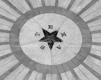 May include: Black and white image of a star-shaped emblem with the letters T, E, X, and A around it. The emblem is centered within a circular design, surrounded by radiating lines. The image has a textured, stone-like appearance.