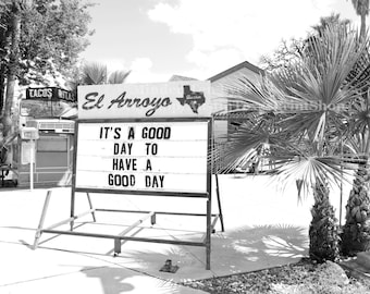 May include: Black and white photo of a sign outside a restaurant called "El Arroyo." The sign reads "It's a good day to have a good day." The sign is in front of a palm tree and a building.
