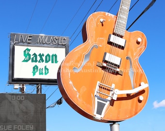 Puede incluir: Una gran escultura de guitarra naranja con una golpeador blanco y cuerdas plateadas. La guitarra está montada en un poste frente a un letrero que dice "Live Music Saxon Pub".