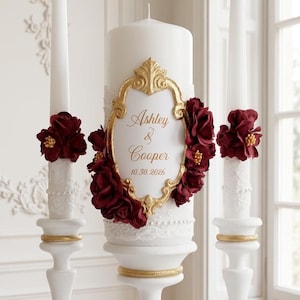 Puede incluir: Un juego de tres velas blancas sobre pedestales blancos. La vela central tipo pilar presenta un marco dorado ornamentado con el texto Ashley & Cooper 10.30.2026, rodeado de rosas de tela rojo oscuro y encaje blanco. Dos velas finas la flanquean.