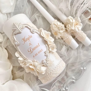 May include: A white wedding candle set. The large pillar candle features a decorative frame with the names "Kayla & Daniel" and the date "11.22.2025". The set includes two taper candles, all adorned with floral and lace accents.