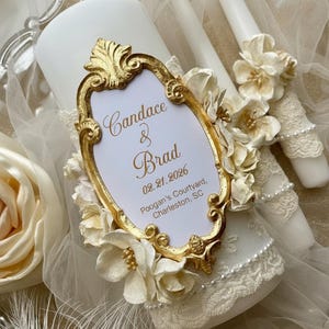 May include: A white pillar candle with a gold-framed label that reads "Candace & Brad 02.21.2026 Poogan's Courtyard, Charleston, SC". The candle is decorated with cream-colored flowers and lace. Two white taper candles are in the background.