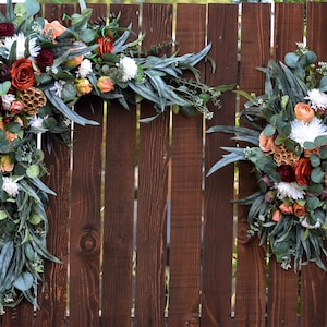 May include: Two floral arrangements with a mix of peach, burgundy, and white flowers, greenery, and dried elements, attached to a brown wooden fence.
