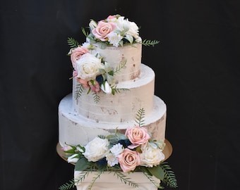 May include: A three-tier wedding cake with a rustic white buttercream frosting and delicate floral decorations. The cake is adorned with pink and white roses, greenery, and blue accents.