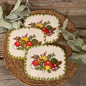 May include: Four oval placemats with a cream background and a green leaf border. Each mat features a colorful illustration of various fruits and vegetables, including apples, mushrooms, and peppers. The mats are arranged on a woven jute mat.