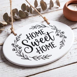 May include: Oval white ceramic sign with the words "HOME Sweet HOME" in black script, surrounded by a floral wreath. The sign hangs from a jute rope. A small terracotta candle holder and eucalyptus sprig are in the background.