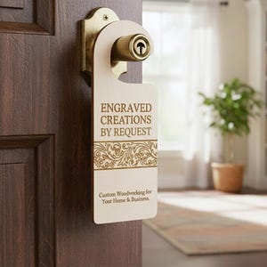 May include: A wooden door hanger with the text "ENGRAVED CREATIONS BY REQUEST" and decorative floral elements. The door hanger is hanging on a door with a brass doorknob. The text "Custom Woodworking for Your Home & Business" is also visible.