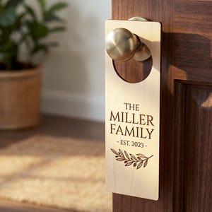 May include: A wooden door hanger with the text "THE MILLER FAMILY - EST. 2023" engraved on it. The door hanger is hanging on a dark brown wooden door. A gold-colored doorknob is visible at the top. A decorative olive branch is at the bottom.