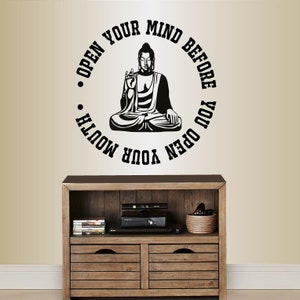 May include: A black and white wall decal featuring a seated Buddha figure within a circular design. The text "OPEN YOUR MIND BEFORE YOU OPEN YOUR MOUTH" surrounds the Buddha. The decal is mounted above a wooden cabinet.