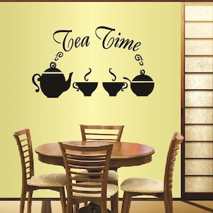 Puede incluir: Una calcomanía de pared en blanco y negro con el texto "Tea Time" e imágenes de una tetera y tazas de té. La calcomanía se muestra en una pared amarilla sobre una mesa de madera con cuatro sillas.