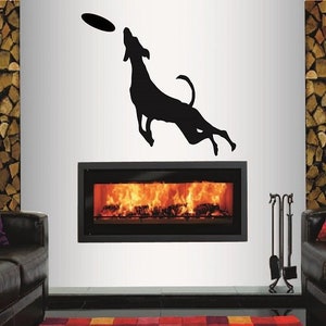 Op de afbeelding: Zwarte silhouet van een hond die springt om een frisbee te vangen, met een open haard op de achtergrond.