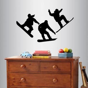 Pode incluir: Adesivo de parede em silhueta preta de três snowboarders sobre um fundo branco. Os snowboarders estão no ar, com suas pranchas viradas para frente. O adesivo é perfeito para o quarto de uma criança ou uma sala de jogos.