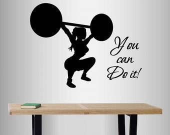 Può includere: Silhouette nera di una donna che solleva un bilanciere sopra la testa in posizione di squat. Il testo "You can Do it!" è scritto in corsivo. L'immagine è su un muro bianco sopra una panca di legno con gambe nere.