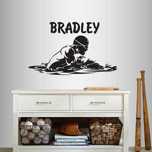 May include: A black and white wall decal featuring a swimmer in action, with the name "BRADLEY" above. The decal is displayed above a white cabinet with baseballs, a glove, and bats.