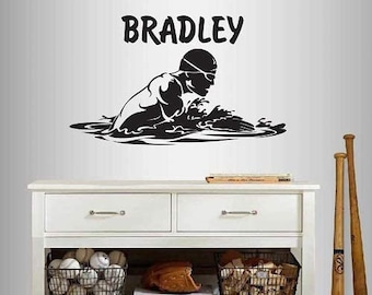 May include: A black and white wall decal featuring a swimmer in action, with the name "BRADLEY" above. The decal is displayed above a white cabinet with baseballs, a glove, and bats.